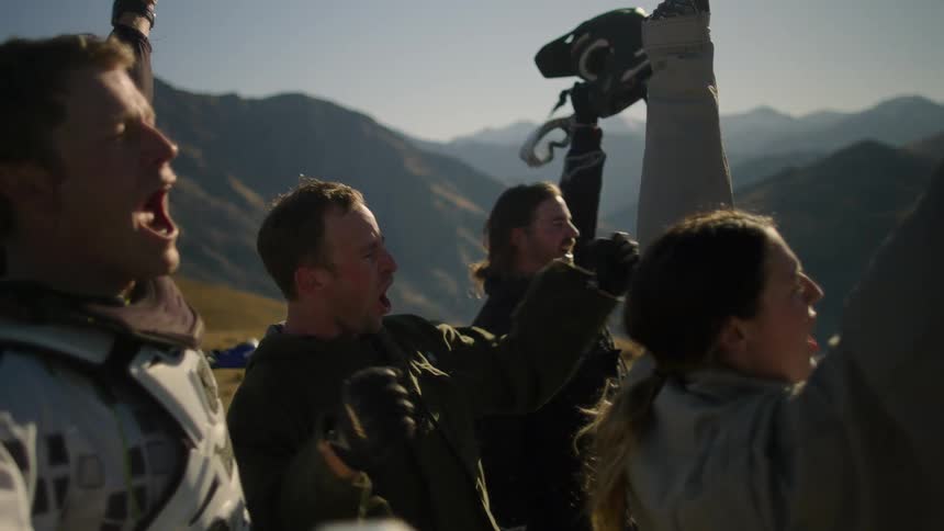 The sun shines upon a small group of people standing in the mountains near motorcycles as they cheer and raise their helmets into the air