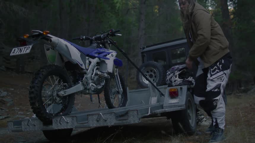 Woman gets out of jeep parked in forest and walks around to free motorcycle on open trailer