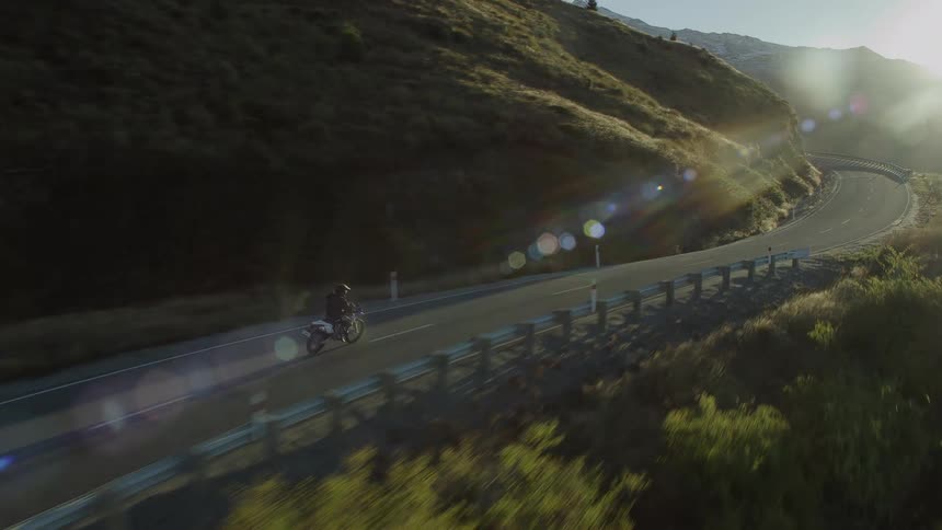 A motorcycle rider races down a twisting highway with green mountains surrounding him