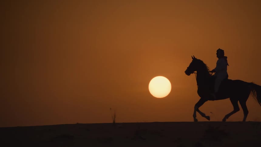 A silhouette of a person riding a horse through the desert as the sun is setting on an orange sky