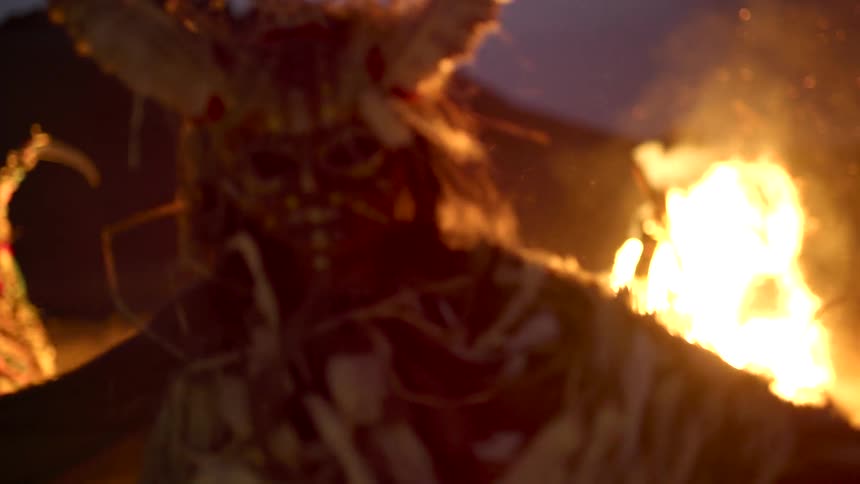 A person wearing a mask and costume dancing around at night around a bonfire on the beach