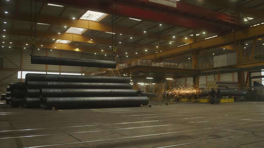 A small group of employees are working in a plant while a large crane positions a long metal culvert