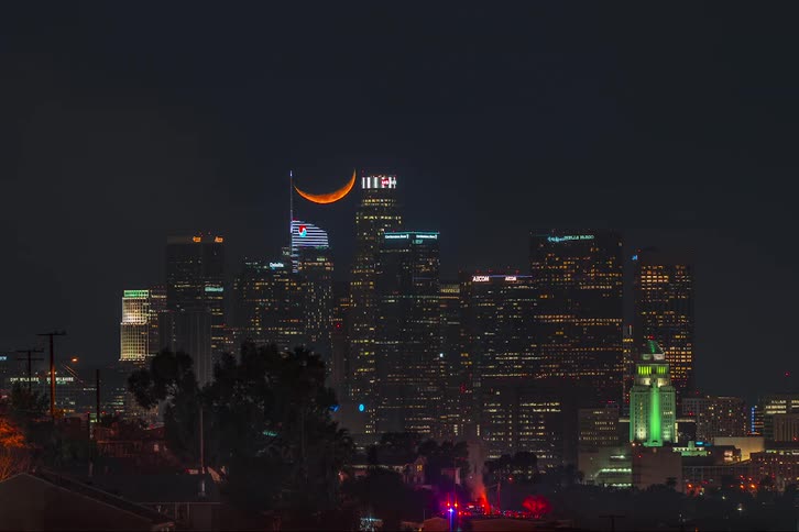 A time lapse of a city at night with a crescent moon lowering over the buildings as a fire erupts in the city and smoke billows into the air