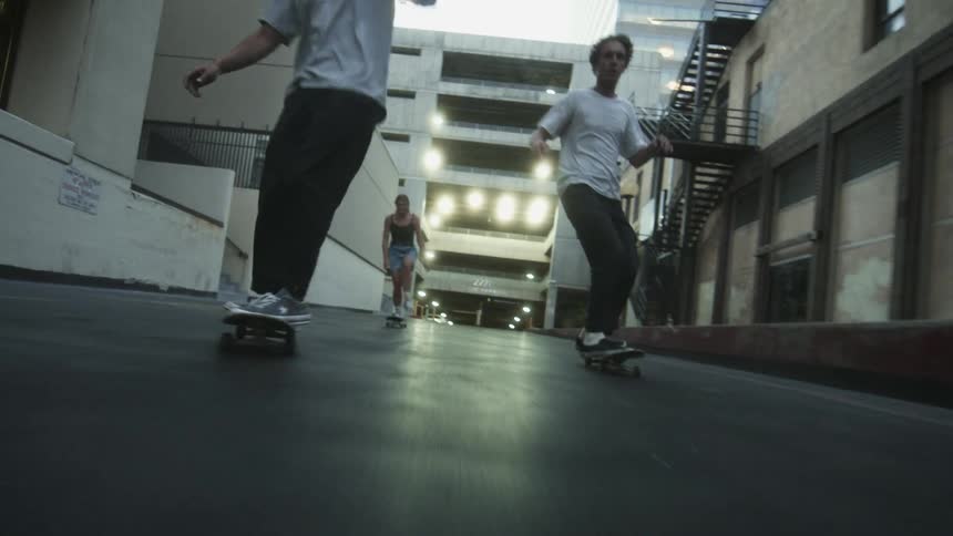 A group of teenagers ride skateboards down an alley past a parking garage
