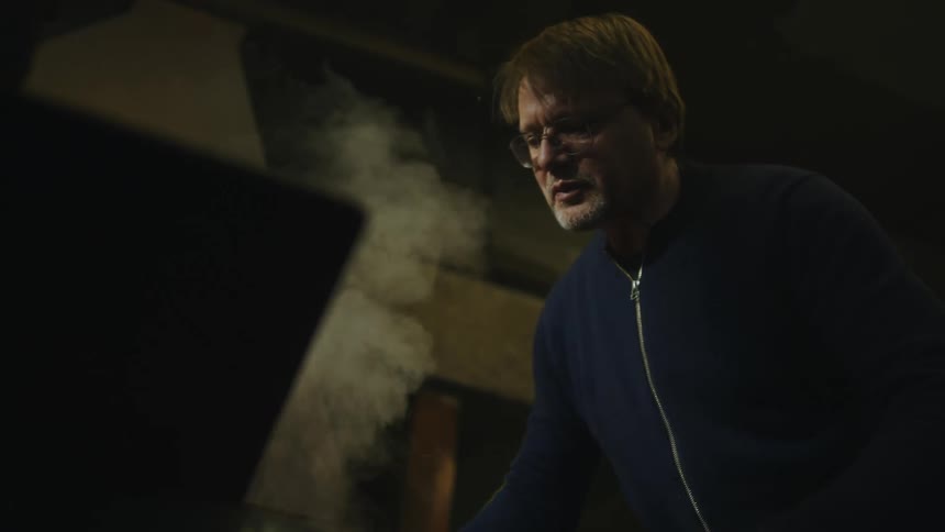 A man wearing glasses leans over something smoking as he lifts a hot metal rod up