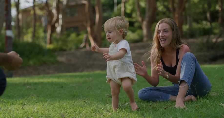 A woman is sitting outside in the grass as her baby girl is learning to walk to daddy