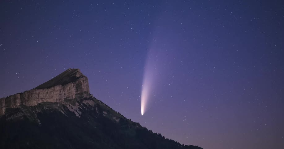 Comet showing tail floats in night sky over forest covered mountain