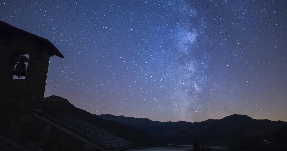 A time lapse of a silhouette of a bell tower and mountains against a purple and blue starry sky