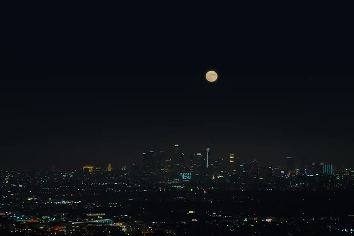 A nighttime view of a city skyline as a full moon rises above the skyscrapers
