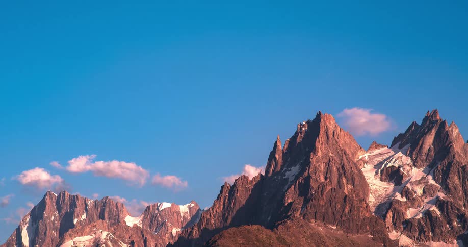 Rough mountain peaks stand high in a blue sky as the moon rises in the distance among a few cumulus clouds