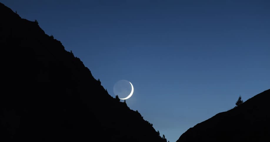 The moon that is a sliver moves down a silhouettelike hill as the sky becomes darker and darker