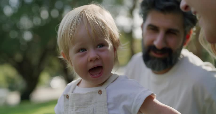 Parents sit with smiling child outdoors as vehicle passes on road in the background