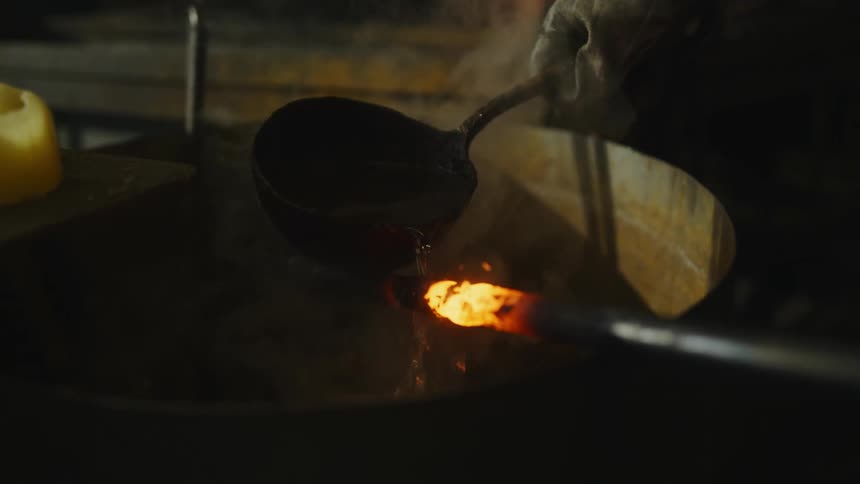 An up close of a man wearing gloves holding a pan full of steaming water and pouring it on a hot iron bar