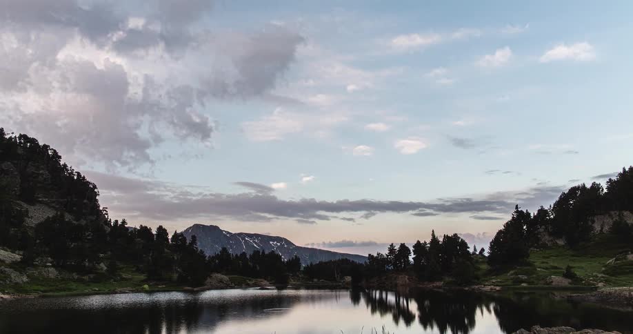 A time lapse of a lake surrounded by forests and mountains in the distance with the sky reflecting on the lake
