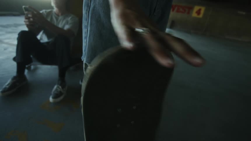 A teenager spins a skateboard on its end inside an urban indoor parking lot while friends sit around