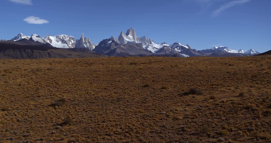 An aerial daytime panorama of flat ground with rocky white mountains in the distance