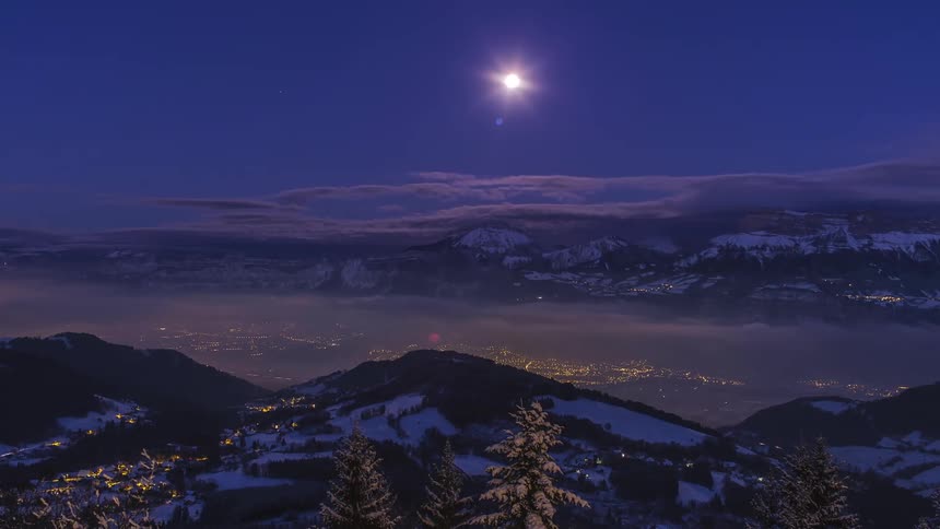 Moon sets and city lights go out as fog flows through valley surrounded by snowy mountains under a brightening sky