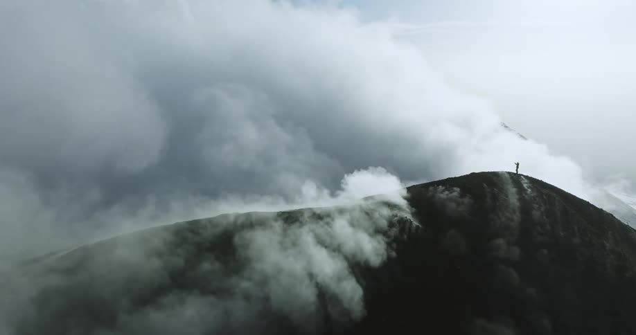 A time lapse of a person standing on top of a mountain in the distance as clouds roll over the mountain top