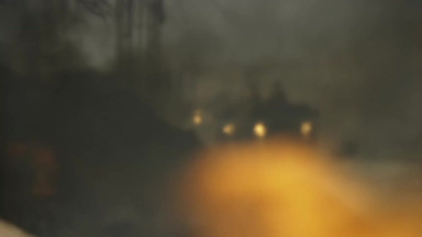 A wrecked car on fire while a military convoy approaches