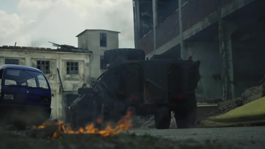 Two military vehicles driving through bombed out area in war zone with fire burning in street