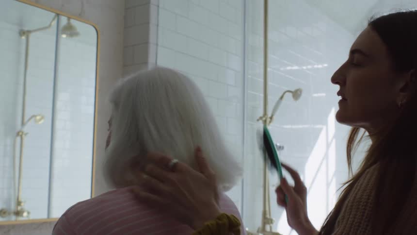 A young woman brushes her grandmas hair in the bathroom mirror