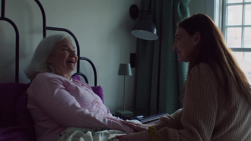 An elderly woman is laying in bed while a younger woman sits next to her holding her hand and talking to her