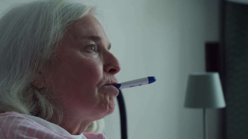 A close up of an older woman sitting in bed having another person stick a thermometer in her mouth as she nods her hod afterwardd