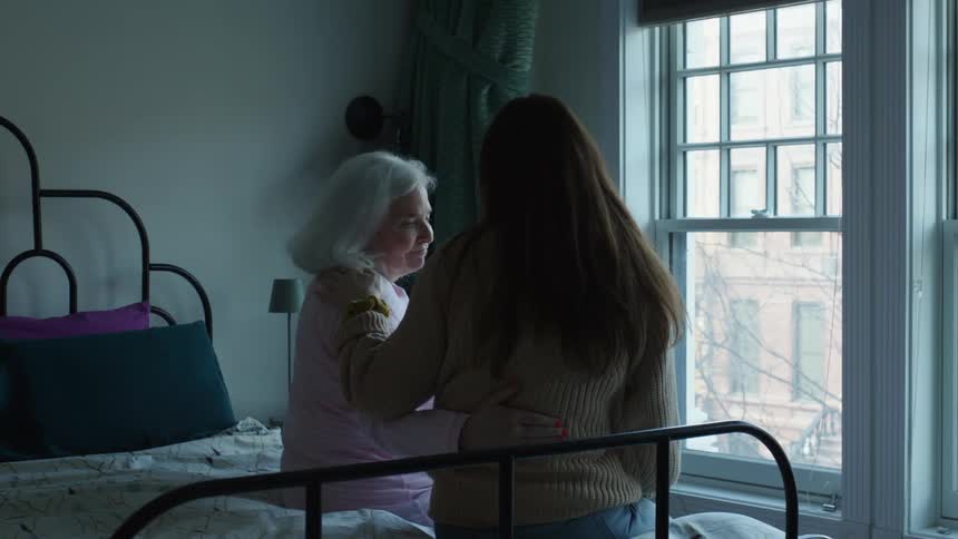 Woman hugs and comforts elderly woman while they sit on bed near windows