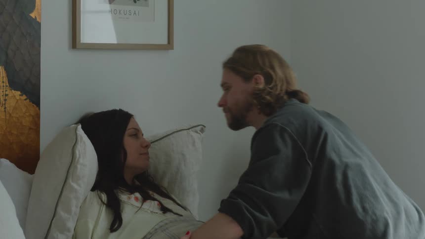A woman lays in bed looking sick while a man sits next to her and kisses her on the head