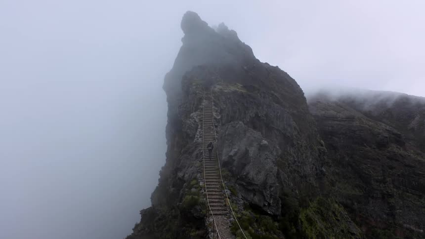 A lone person crosses a wooden bridge high up on a hazy mountainous cliff
