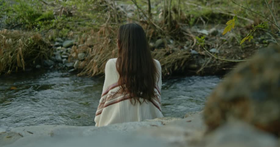 The backside of a woman sitting next to a flowing creek