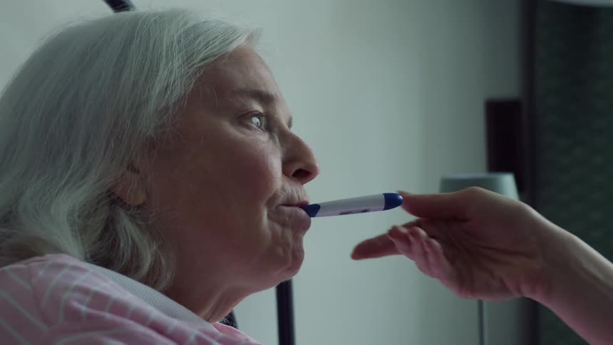 Younger woman checks temperature of elderly female relative lying in bed