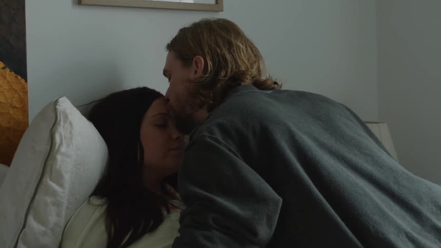 A man tending to a sick woman in a bed stroking her hair and giving her a kiss on the forehead