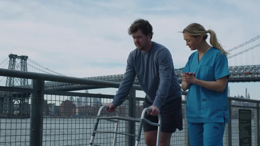 Woman wearing scrubs encourages man using walker as they move along waterfront with bridge and city in background