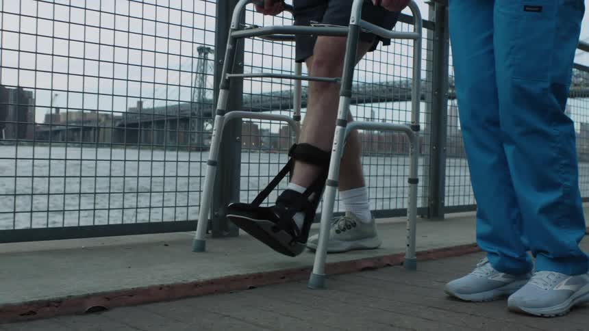 A nurse walks next to a man with his foot in a brace as he uses a walker