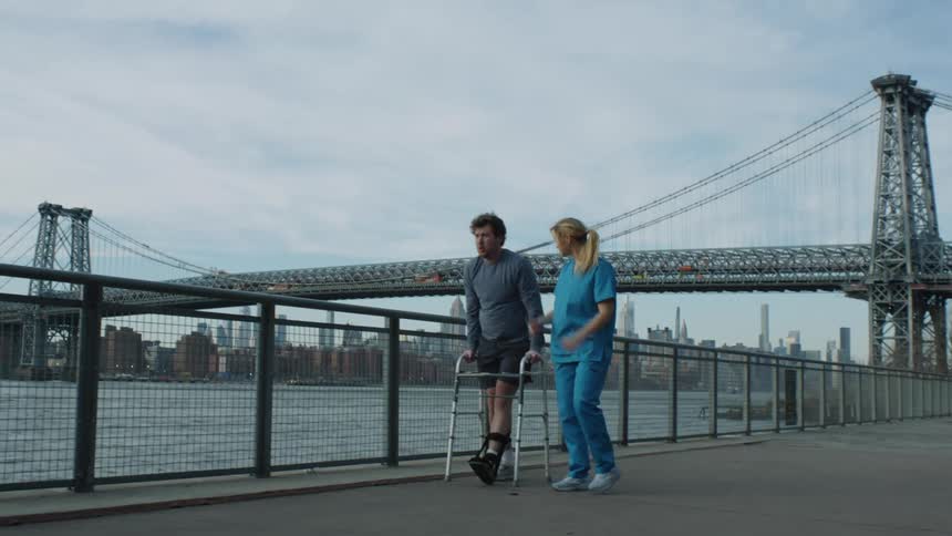 A nurse encourages a man in a leg brace as he walks along a waterfront using a walker