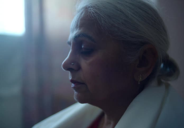 An elderly woman sits calmly indoors near a sunlight window