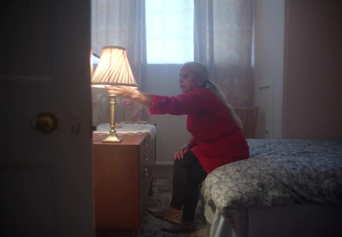 Elderly woman with red shirt and black pants sits on bed and turns off bedside lamp in bedroom