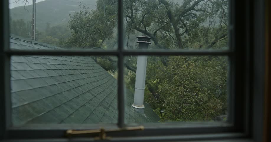 A chimney on the roof of a house is shown through the perspective of inside of another house looking through a window