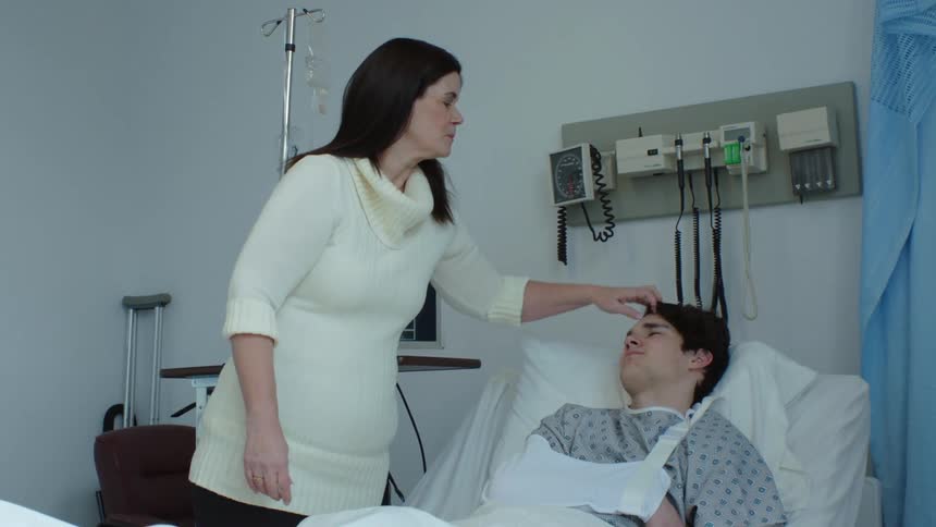 A teenage boy in a hospital bed as a patient with a sling on his arm and his mother pushing his hair back
