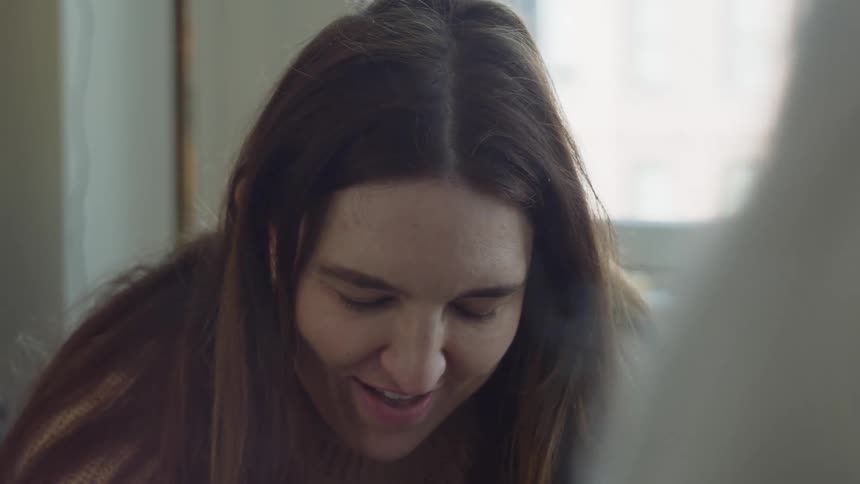 A close up of a woman wearing a tan sweater looking down laughing and smiling
