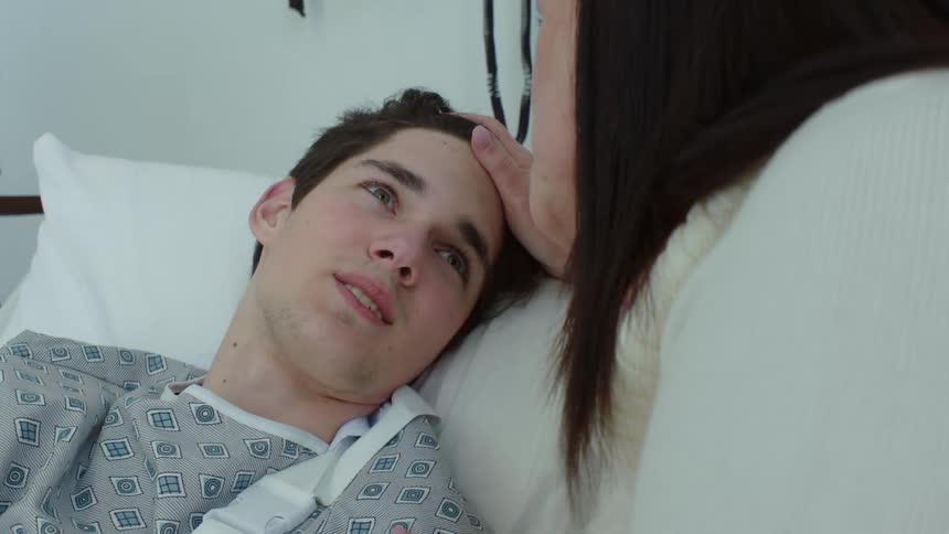 A teenage boy is lying in a hospital bed as his mothers visits and has her hand on his head as she speaks to him