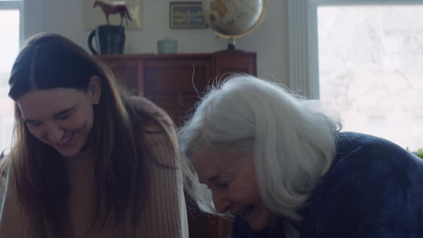 An older woman and a younger woman sit on the floor laughing and smiling while doing something on the floor