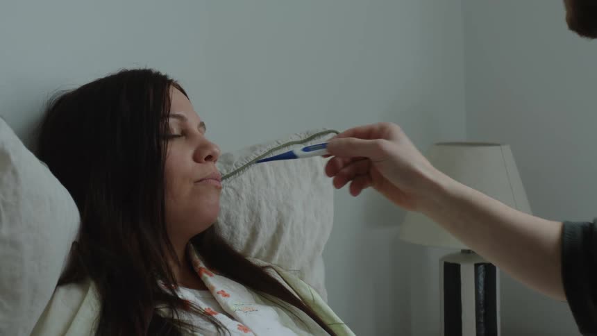 Young man puts a thermometer on his sick wife mouth while she lays in bed at home