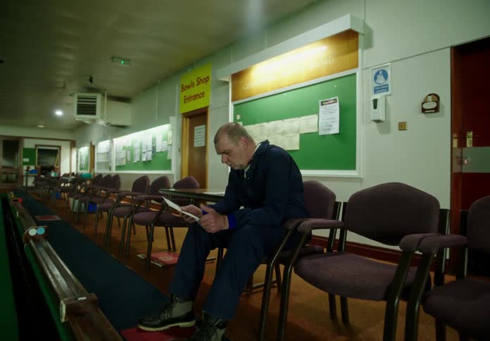 A man is sitting alone in an indoor golf range shaking as he holds a sheet of paper and reads it