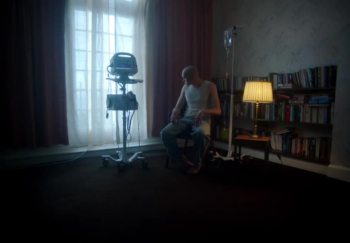 A depressed cancer patient sits in a darkened room near medical equipment while glancing outdoors