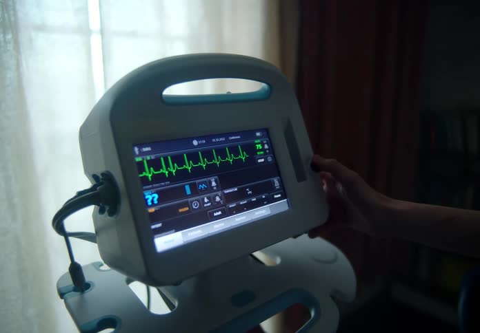 A human hand touches an active medical heart rate monitor near a window in a darkened room