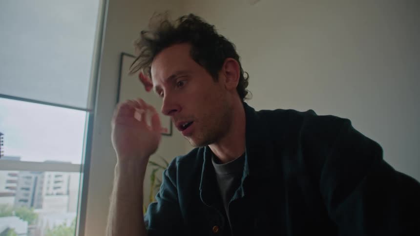 A man is sitting on the couch in front of a table typing on a laptop computer and he stops to rub his head