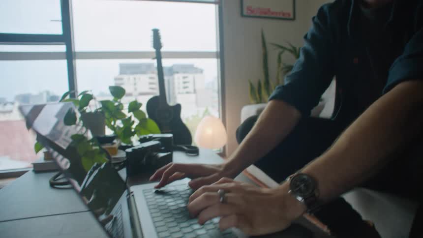 A man is sitting on the couch in front of a table typing on a laptop computer and he stops to think