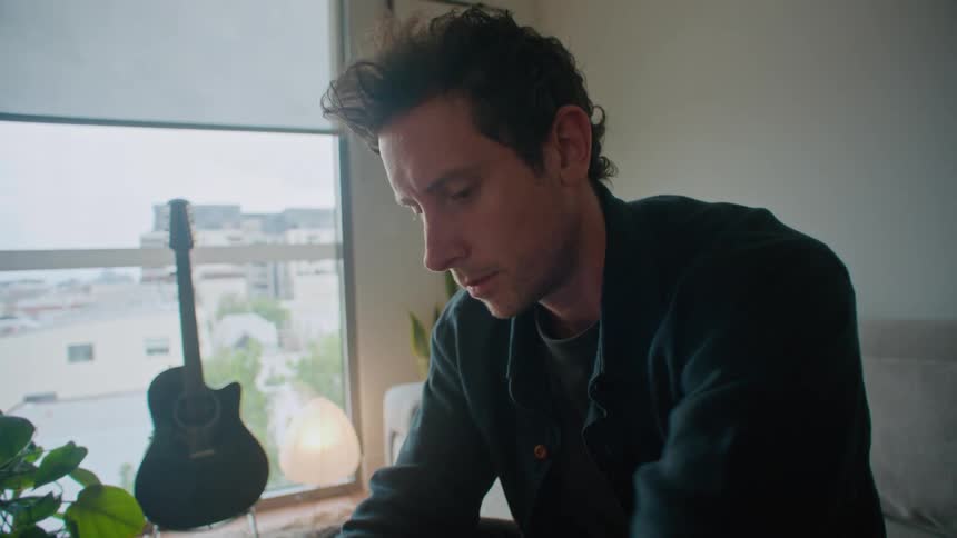 A man is typing on his laptop and looking at a camera